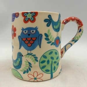 May include: A ceramic mug with a whimsical design featuring blue owls, birds, flowers, and trees on a cream background. The handle is decorated with red and white accents. The mug is a standard size for hot beverages.