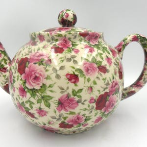 Peut inclure: Théière en céramique crème ornée d'un motif floral de roses roses et bordeaux et de feuilles vertes. La poignée, le bec et le couvercle présentent le même motif. La théière a une forme arrondie, idéale pour servir le thé.