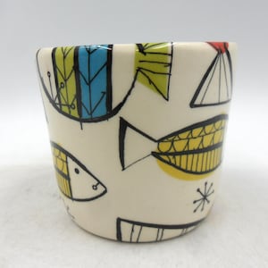May include: A ceramic cup with a cream background, decorated with a repeating pattern of stylized fish in yellow, blue, and green. The fish are outlined in black, with red accents. The cup has a slightly flared shape.