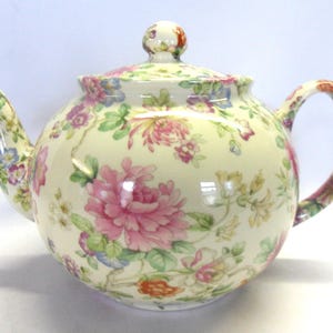 May include: A cream-coloured teapot with a floral design. The teapot has a round body, a lid with a small knob, a curved handle, and a spout. The floral pattern includes pink, purple, and blue flowers with green leaves. The teapot is suitable for serving tea.