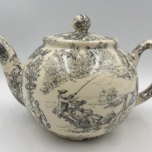 May include: A white ceramic teapot with a black and white toile pattern depicting a pastoral scene. The teapot has a round body, a spout, a handle, and a lid.