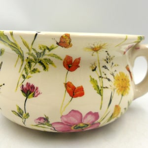 May include: A cream-coloured ceramic chamber pot featuring a colourful botanical print of various wildflowers, including red poppies, yellow dandelions, and pink blossoms, along with a small orange butterfly and a sturdy side handle.
