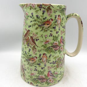 May include: A ceramic jug with a light green background and a pattern of birds, flowers, and foliage. The jug has a large handle and a spout for pouring. The birds are brown, red, and yellow.