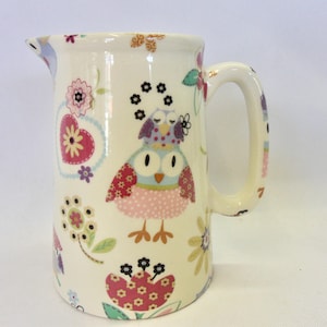 Könnte beinhalten: Eine weiße Keramikkanne mit Henkel, verziert mit einem bunten Muster aus Eulen, Herzen und Blumen. Die Eulen sind rosa, blau und lila mit schwarzen Augen. Die Herzen sind rot mit weißen und gelben Blumenmustern. Die Blumen sind rosa, gelb und grün.