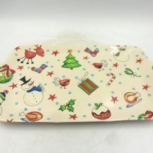 Peut inclure: Plat à servir rectangulaire en céramique avec un fond crème et des illustrations sur le thème de Noël. Le plat présente des dessins de bonhomme de neige, de rennes, de sapin de Noël et d'autres éléments festifs.