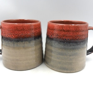May include: Two ceramic mugs featuring a gradient glaze that transitions from a textured, vibrant red at the rim to a dark charcoal middle section, ending in a smooth, light beige base. Each mug has a sturdy, curved handle and a cylindrical body.