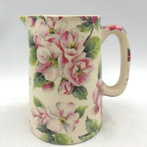May include: Cream ceramic jug with a floral pattern. The jug has a handle and spout, and is decorated with pink and white flowers and green leaves. The floral design covers the entire surface of the jug, a classic kitchenware item.