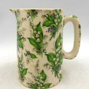 Peut inclure: Une cruche en céramique vintage avec un fond crème et un motif floral de feuilles vertes et de fleurs blanches et violettes. La cruche a une anse et un bec verseur.
