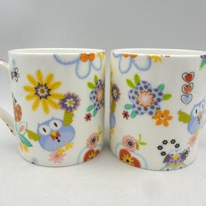 May include: Two white ceramic mugs featuring a floral and owl design. The mugs have a white background with colourful flowers, owls, and hearts.