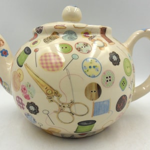 May include: A white ceramic teapot decorated with a colourful design of buttons, thread, scissors and other sewing-related items.