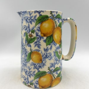 Könnte beinhalten: Hoher Keramik-Krug mit blauem Blumenmuster und gelben Zitronen auf weißem Grund. Der Krug hat einen Griff und einen Ausguss. Das Design umfasst grüne Blätter und kleine weiße Blüten. Ein dekorativer Küchenartikel.
