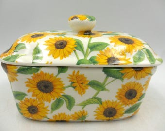 Puede incluir: Un recipiente de mantequilla de cerámica blanca con tapa. El recipiente y la tapa están decorados con un patrón de girasoles amarillos sobre un fondo blanco.