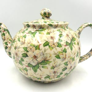Peut inclure: Une théière en céramique blanche avec un motif floral de fleurs blanches et de feuilles vertes. La théière a un couvercle et une poignée.