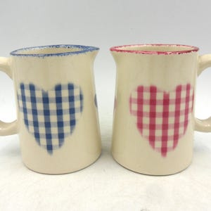 May include: Two ceramic creamers with handles. One has a blue and white gingham heart design and the other has a pink and white gingham heart design.