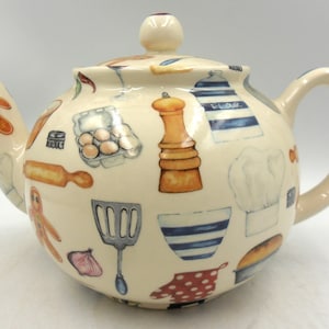 May include: Cream-coloured ceramic teapot decorated with kitchen-themed illustrations. The teapot features images of a gingerbread man, eggs, a rolling pin, a chef's hat, and a pepper grinder. The handle and spout are a matching cream colour.