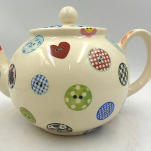 May include: Cream ceramic teapot adorned with colourful button designs. The teapot has a rounded body, a curved spout, and a loop handle. The buttons are in various colours and patterns, including red hearts and checkered designs.