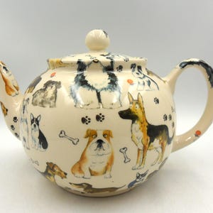 May include: Cream-coloured ceramic teapot decorated with various dog breeds, paw prints, and bones. The teapot has a rounded body, a curved handle, and a spout. The lid is topped with a small, round knob.