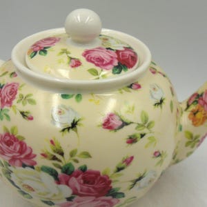 May include: A cream-colored teapot with a floral pattern of pink and white roses. The teapot has a lid with a knob on top.