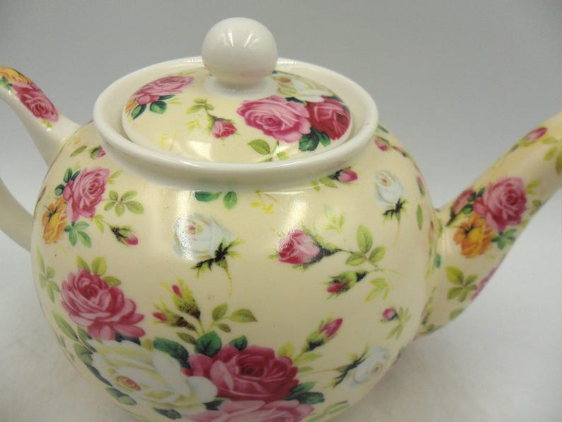 May include: A cream-colored teapot with a floral pattern of pink and white roses. The teapot has a lid with a knob on top.