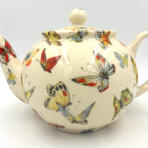 Peut inclure: Une théière en céramique blanche avec un motif de papillon. La théière a un corps rond et un bec courbé. La poignée est sur le côté droit de la théière. La théière est décorée de papillons colorés dans différentes nuances de rouge, jaune, bleu et vert.