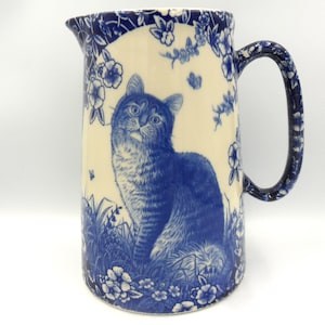 May include: A blue and white ceramic jug with a cat design. The cat is sitting in a field of flowers and is looking up. The jug has a handle and a spout.