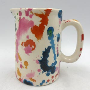 May include: A white ceramic jug with a colourful splatter paint design. The jug has a handle and is approximately 15 cm tall.