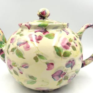 May include: A white ceramic teapot with a floral pattern of pink and purple sweet peas on a cream background. The teapot has a lid with a knob and a handle.