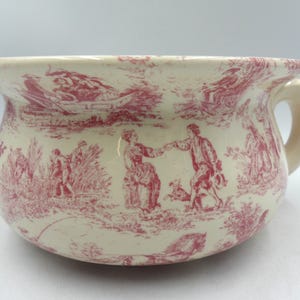 May include: Cream ceramic chamber pot with a red toile de Jouy pattern. The pot has a rounded shape, a handle, and a flared rim. The design shows figures in a rural scene.