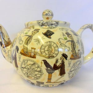 Peut inclure: Théière en céramique crème avec un motif fantaisiste représentant des squelettes, des horloges et d'autres éléments gothiques. La théière a un corps arrondi, une anse incurvée et un bec verseur avec une figurine de squelette.