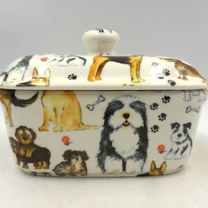 May include: White ceramic butter dish with a lid. The dish is decorated with a pattern of dogs of various breeds, including a sheepdog, a terrier, and a dachshund. The dogs are depicted in a cartoon style and are surrounded by paw prints and bones.