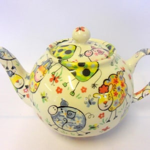 May include: A white ceramic teapot with a whimsical design featuring colourful cartoon animals and flowers. The teapot has a round body and a lid with a knob in the shape of a flower.