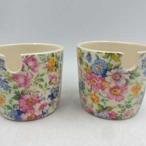 May include: Two white ceramic sugar bowls with a floral pattern in shades of pink, blue, yellow, and green. Each bowl has a cut-out on the side for easy pouring.