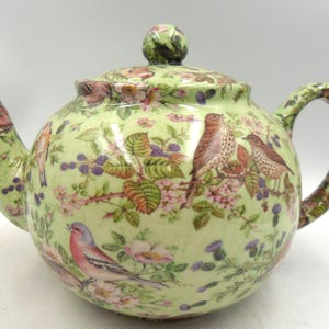 May include: A round, pale green ceramic teapot with a lid, spout, and handle. The teapot is decorated with images of birds, flowers, and berries. The handle and spout are also decorated with the same pattern. The lid has a small, round knob.