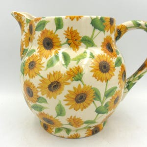 May include: A white ceramic jug with a sunflower pattern. The jug has a round body and a handle. The sunflowers are yellow with brown centres and green leaves.