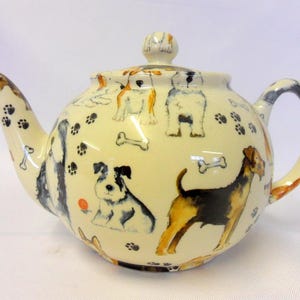 May include: A white ceramic teapot with a dog-themed design. The teapot features a variety of dog breeds, including a terrier, a dachshund, and a poodle. The teapot is decorated with paw prints, bones, and a red ball.