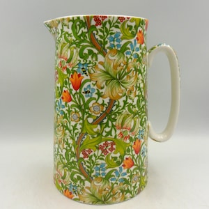 May include: A white ceramic jug with a floral pattern in shades of green, orange, yellow, and blue. The jug has a handle and a wide mouth.