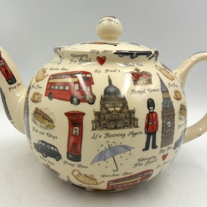 May include: Cream-coloured ceramic teapot adorned with London-themed illustrations. Depicts a red double-decker bus, Big Ben, a red post box, and the text "It's Raining Again". Includes a lid and a curved handle.