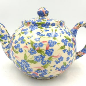 May include: A white teapot with a floral pattern of blue forget-me-nots and pink ribbons. The teapot has a lid and a spout.