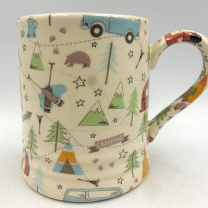 May include: A white ceramic mug with a camping theme. The mug features a variety of illustrations, including a car, a bear, a tent, trees, and mountains. The text "Camping" is printed on a banner.