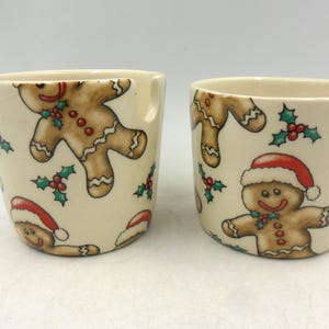 May include: Two white ceramic mugs with a Christmas gingerbread man design. The mugs have a cream background with red, green, and brown gingerbread men.