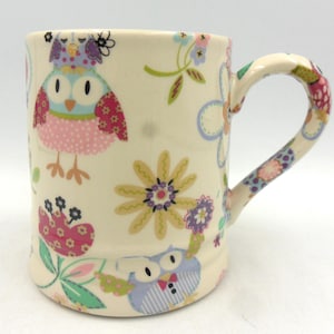 May include: A white ceramic mug with a handle, featuring a colourful owl and floral pattern. The owl is pink, blue, and green with a white background. The flowers are yellow, pink, and purple.