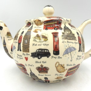 May include: A cream-coloured ceramic teapot featuring London landmarks such as Big Ben, a red bus, and a black cab. The teapot has a rounded body, a curved handle, and a spout, with various illustrations around the body.