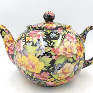Könnte beinhalten: Keramik-Teekanne mit schwarzem Hintergrund und floralem Muster in Gelb, Rosa und Blau. Die Teekanne hat einen runden Körper, einen gebogenen Griff und einen Ausguss. Der Deckel ist mit einem kleinen, runden Knopf versehen.