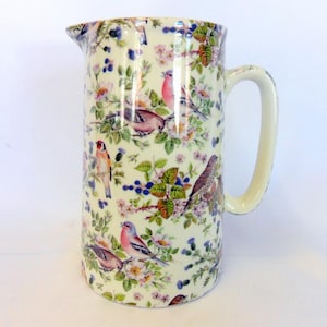 May include: A white ceramic jug with a handle, featuring a floral pattern with colourful birds. The jug has a cream background with a variety of flowers and birds in shades of pink, blue, green, and yellow.