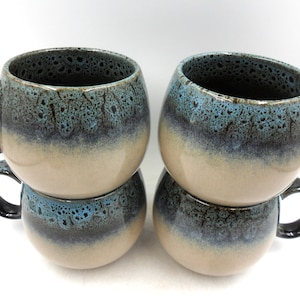 Peut inclure: Quatre tasses en céramique empilées par paires. Les tasses ont une forme arrondie avec une anse marron foncé. L'extérieur présente un dégradé de couleurs, commençant par une base beige, passant au bleu et au marron foncé en haut.