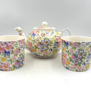 May include: A floral tea set comprising a teapot and two mugs. The set is adorned with a pattern of pink, blue, and yellow flowers on a white backdrop. The teapot includes a lid and a curved spout.