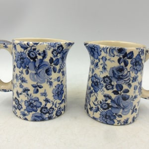 May include: Two ceramic jugs with a cream base and a blue floral pattern. Each jug has a handle and a spout. The floral design features roses and other flowers.
