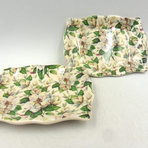 Könnte beinhalten: Zwei weiße Keramikteller mit einem Blumenmuster aus weißen Blüten und grünen Blättern. Die Teller haben einen gewellten Rand.