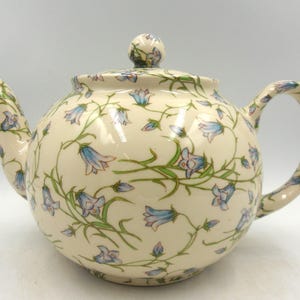 May include: A white ceramic teapot with a floral pattern of blue and green bellflowers. The teapot has a round body and a curved spout.