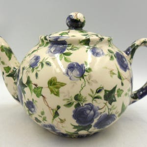 May include: A cream-coloured ceramic teapot featuring a floral design of blue roses and green ivy. The teapot has a rounded body, a curved spout, and a loop handle. The lid is topped with a small, round knob.